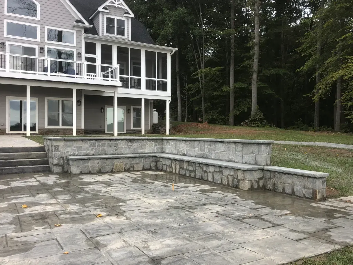 Stone patio with built-in seating beside a gray house and wooded yard