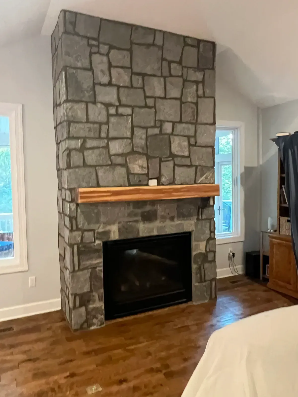 Stone fireplace with wooden mantel in a bright room, beside windows and hardwood floors