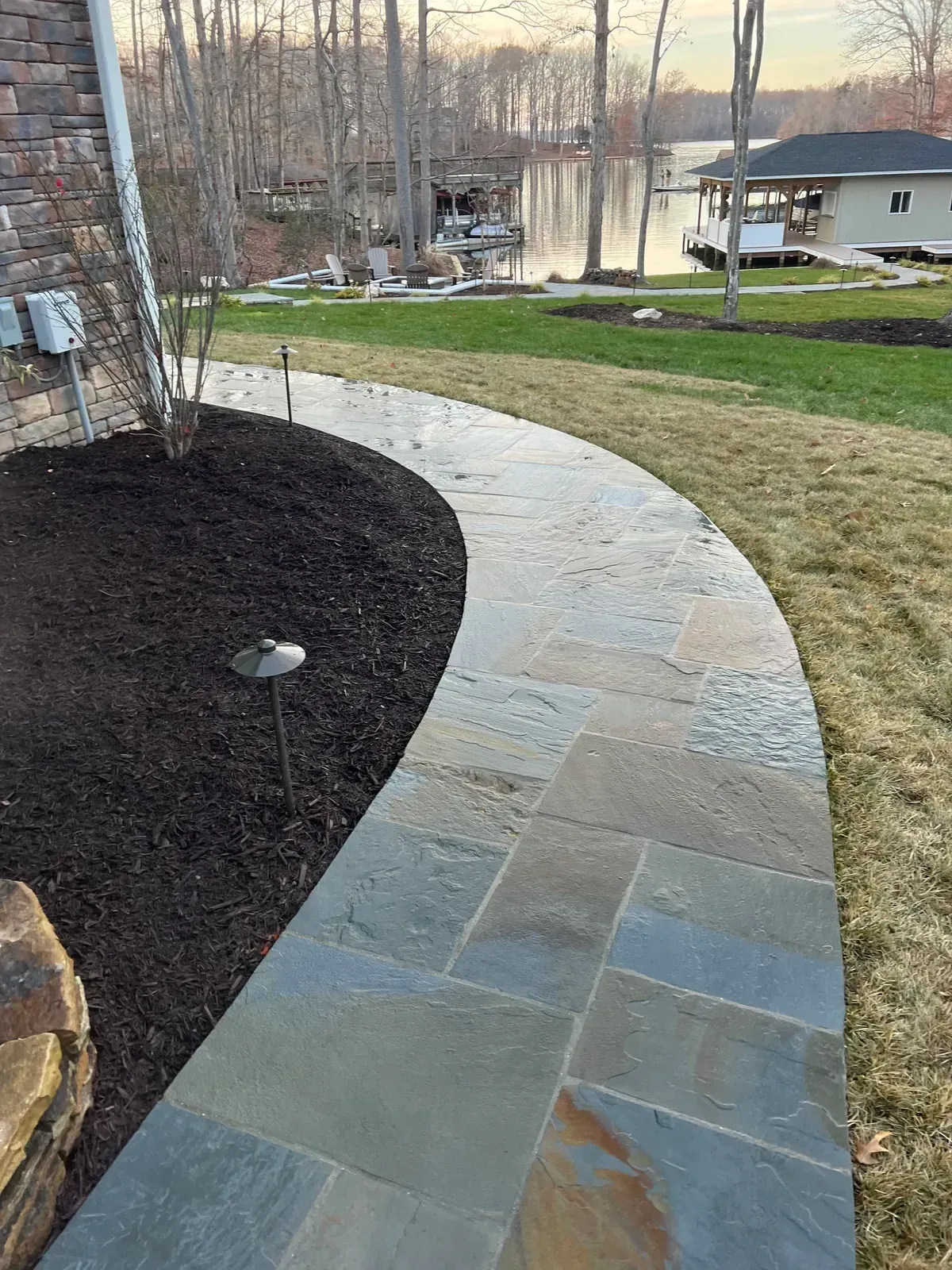 Curved stone walkway beside mulch and lawn, leading toward a lakeside house at dusk