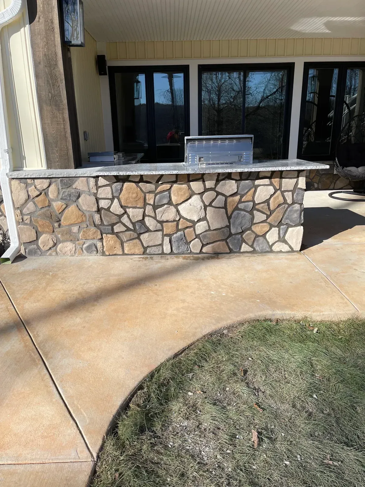 Stone outdoor kitchen counter on a patio with glass doors and a curved concrete walkway in front