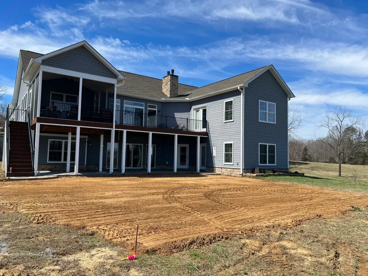 Gray two-story house under construction with wraparound porch and dirt yard