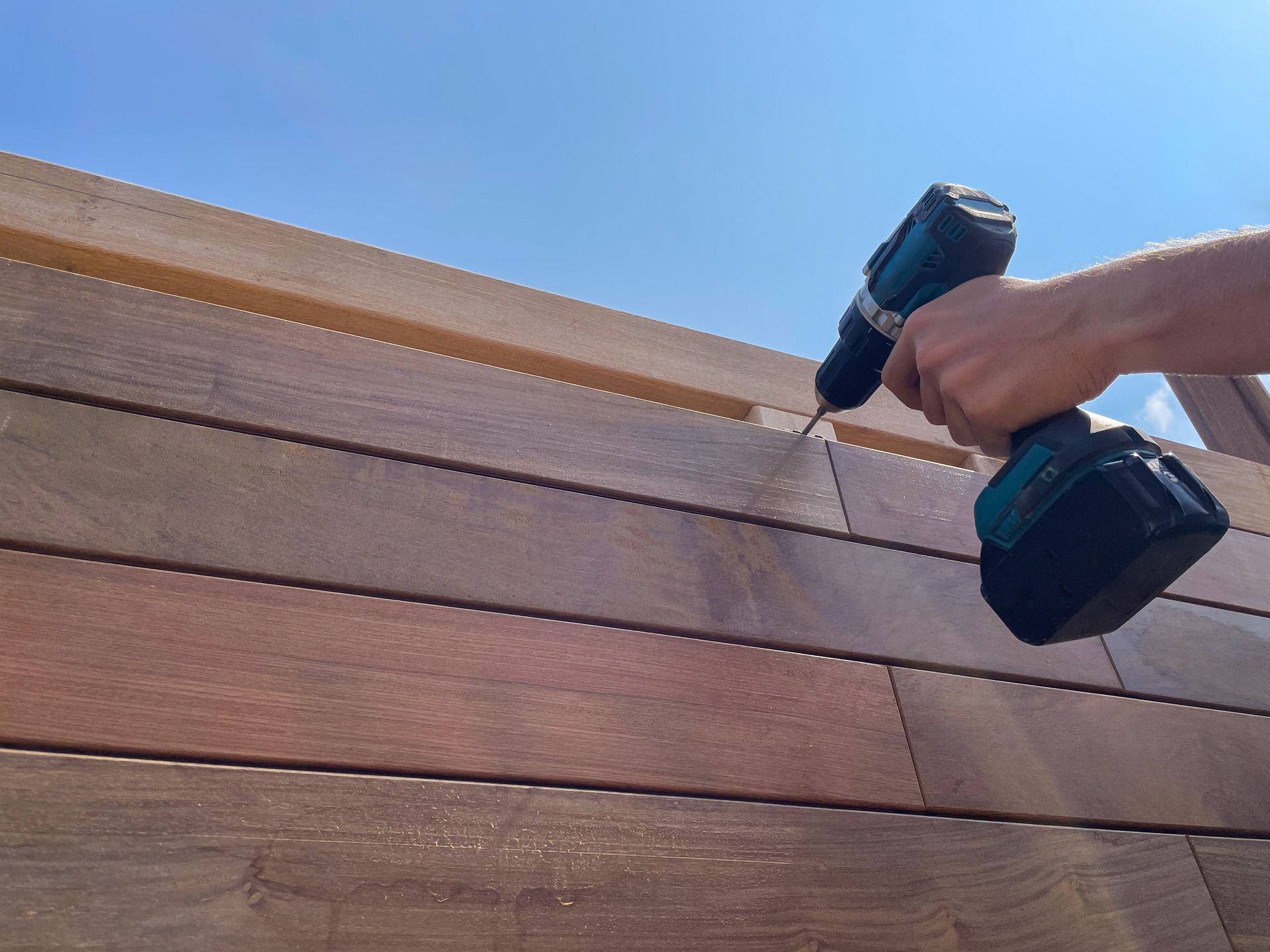 Person using a drill to fasten a screw into a wooden fence, blue sky in the background.