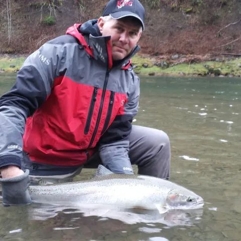 A man in a simms jacket is kneeling in the water holding a fish