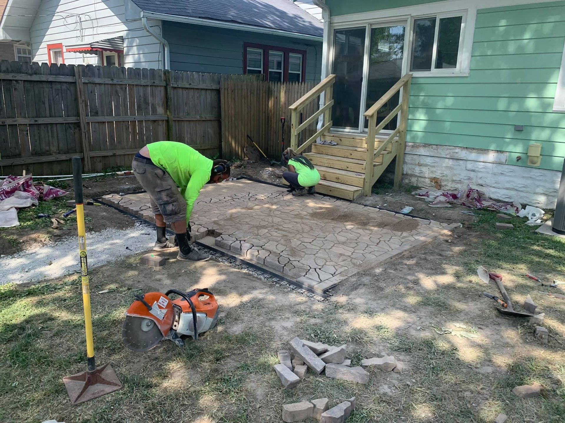 Two workers in neon green shirts repairing a cracked backyard patio beside a house.