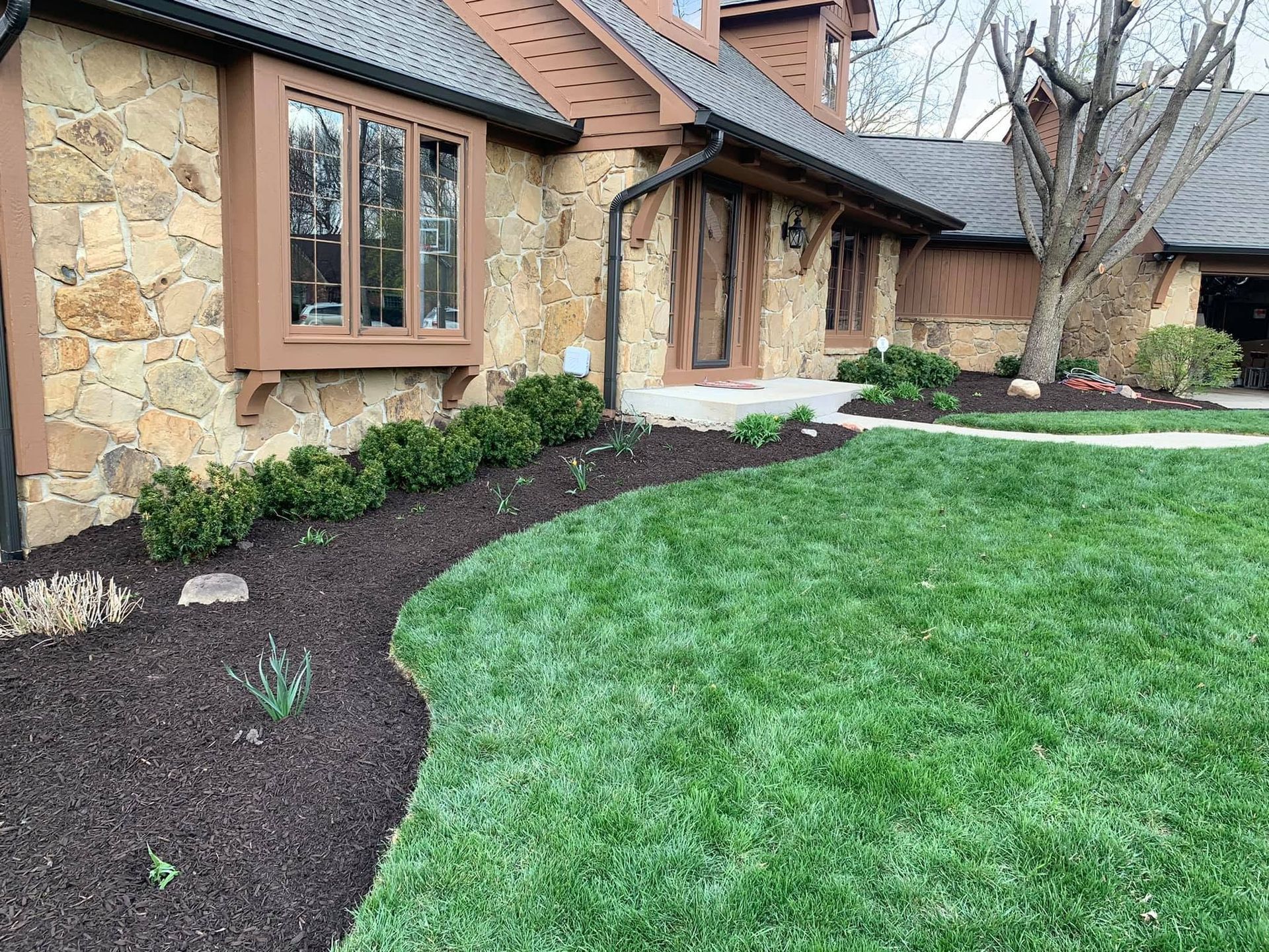 Front yard with green lawn, mulched flower beds, and a stone-and-stucco house with windows.