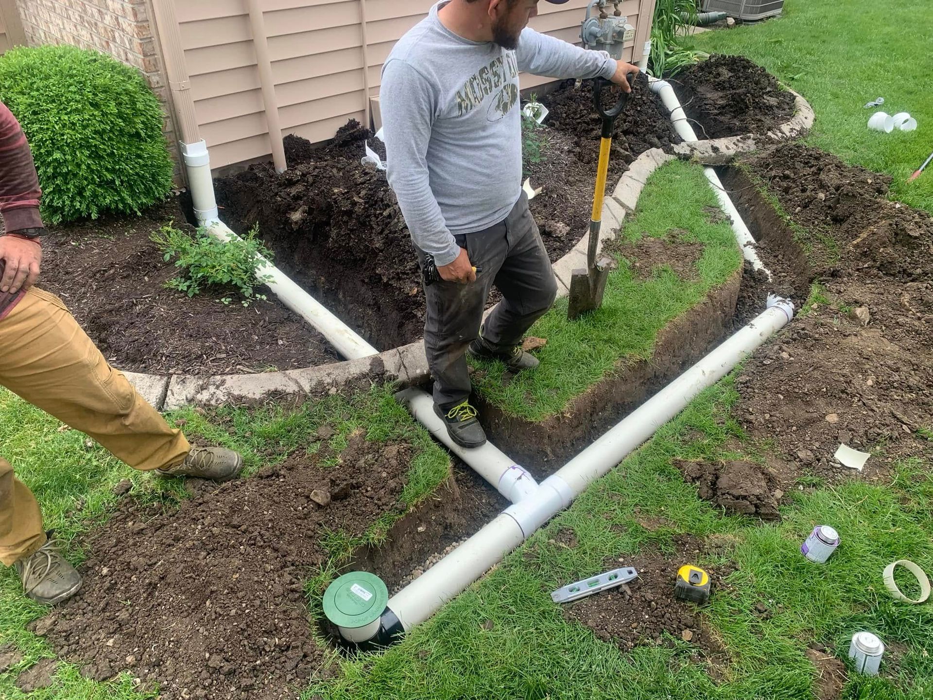 Workers installing a white PVC drain pipe in a muddy yard beside a house.