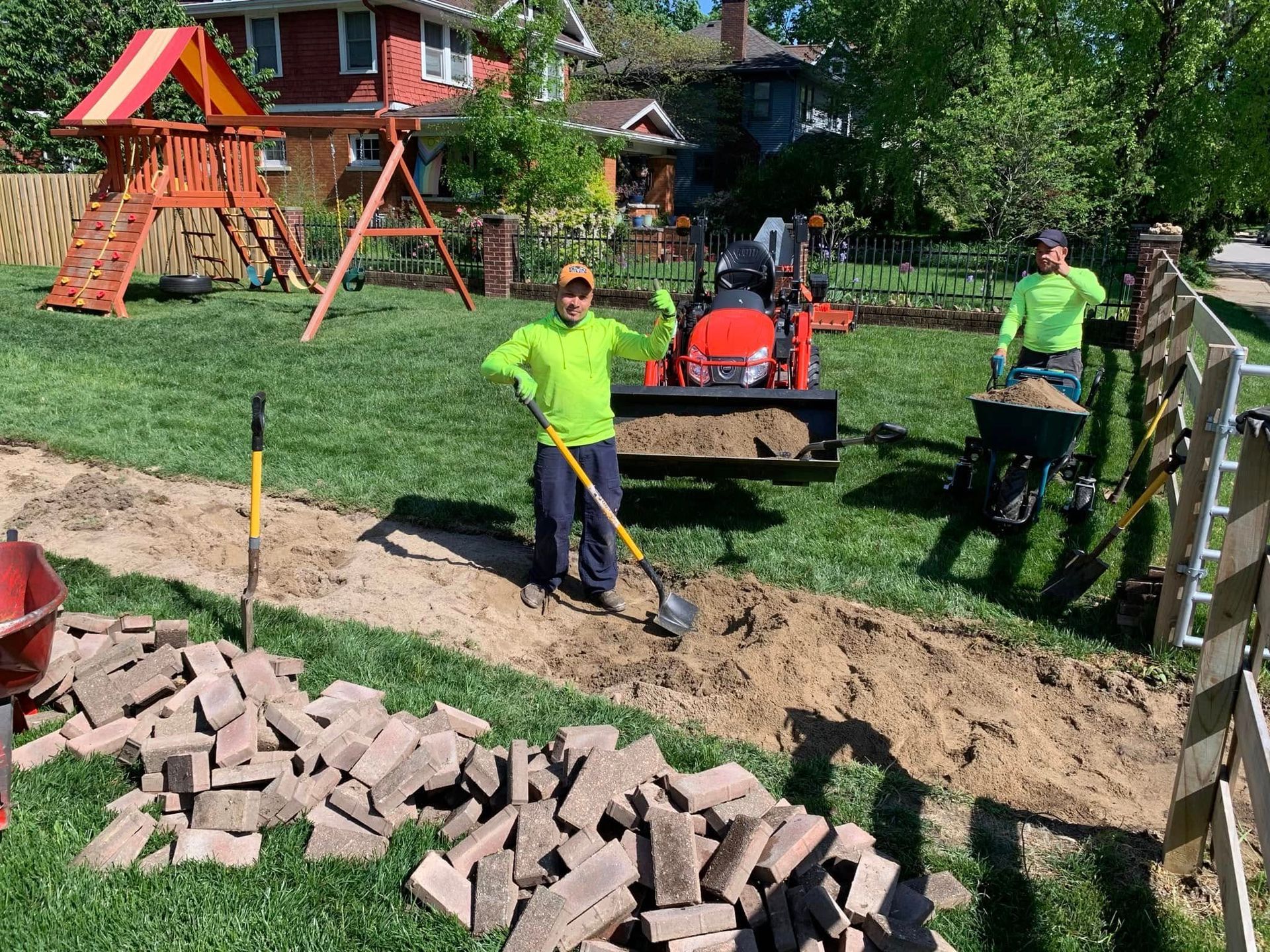 Workers in neon shirts landscaping a yard with a red excavator and stacked pavers