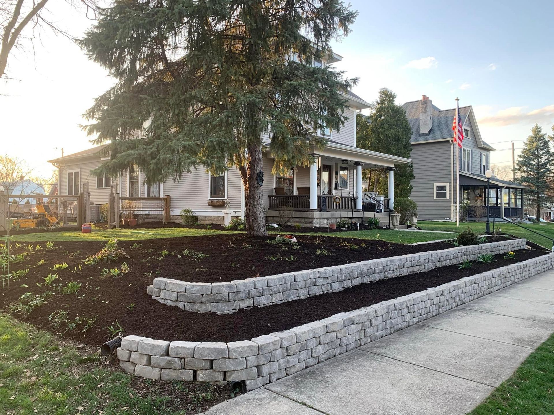 Residential house with front porch, large tree, and landscaped retaining wall beside a sidewalk at sunset