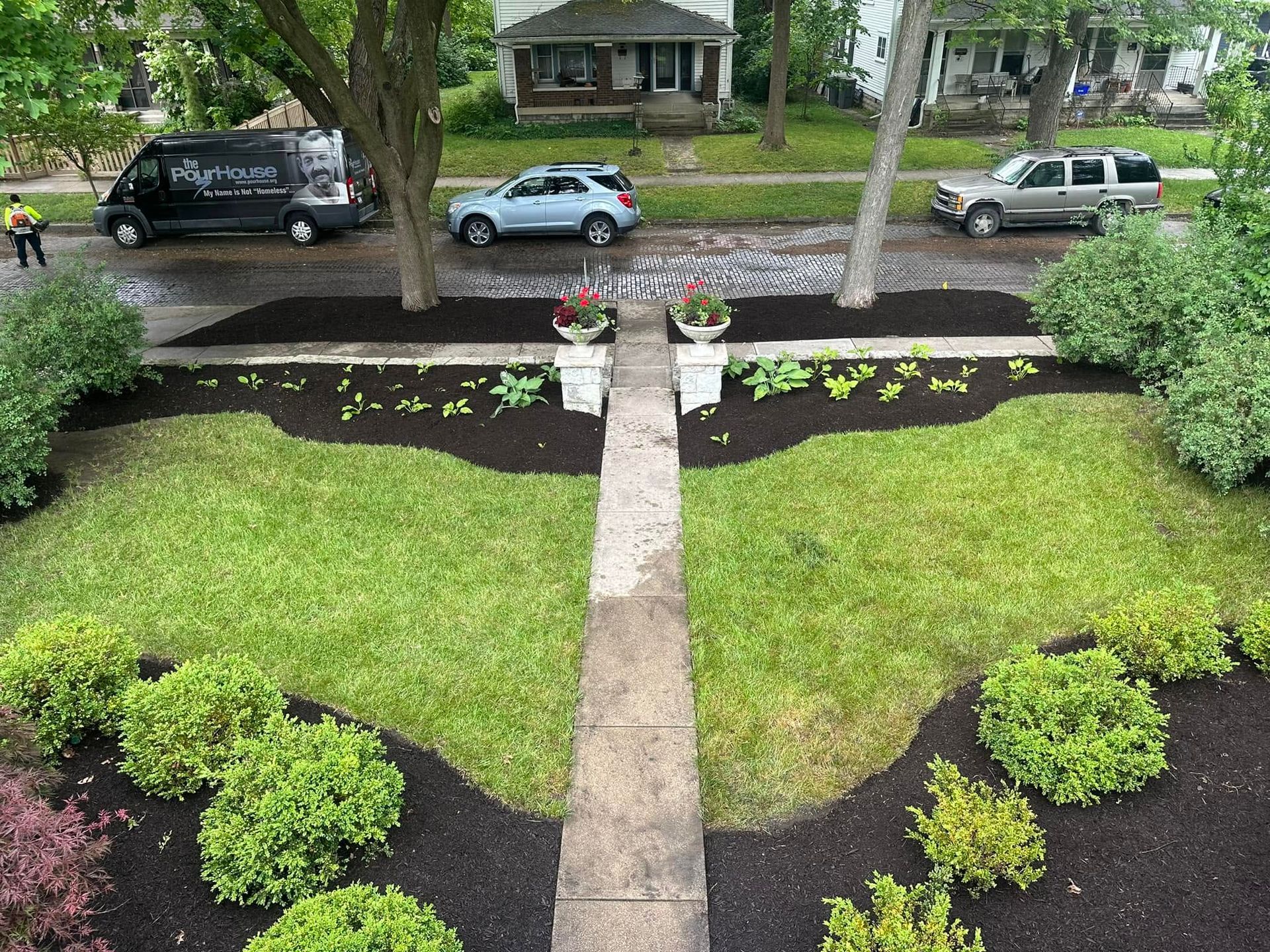 Front yard with a concrete path, green lawn, dark mulch beds, shrubs, and potted plants by the street.