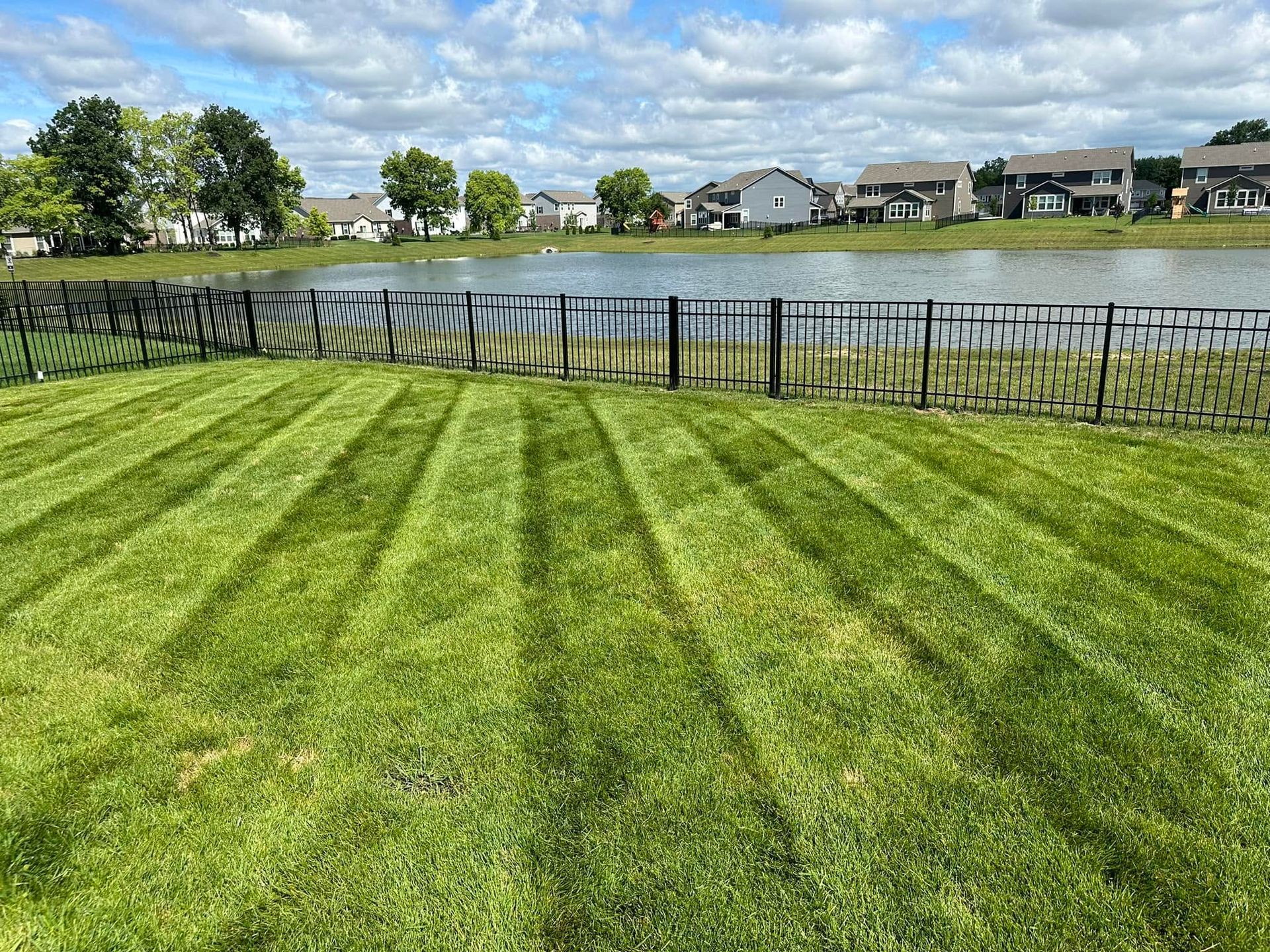 Grassy park with striped mowing lines, black fence, lake, and houses under a cloudy sky