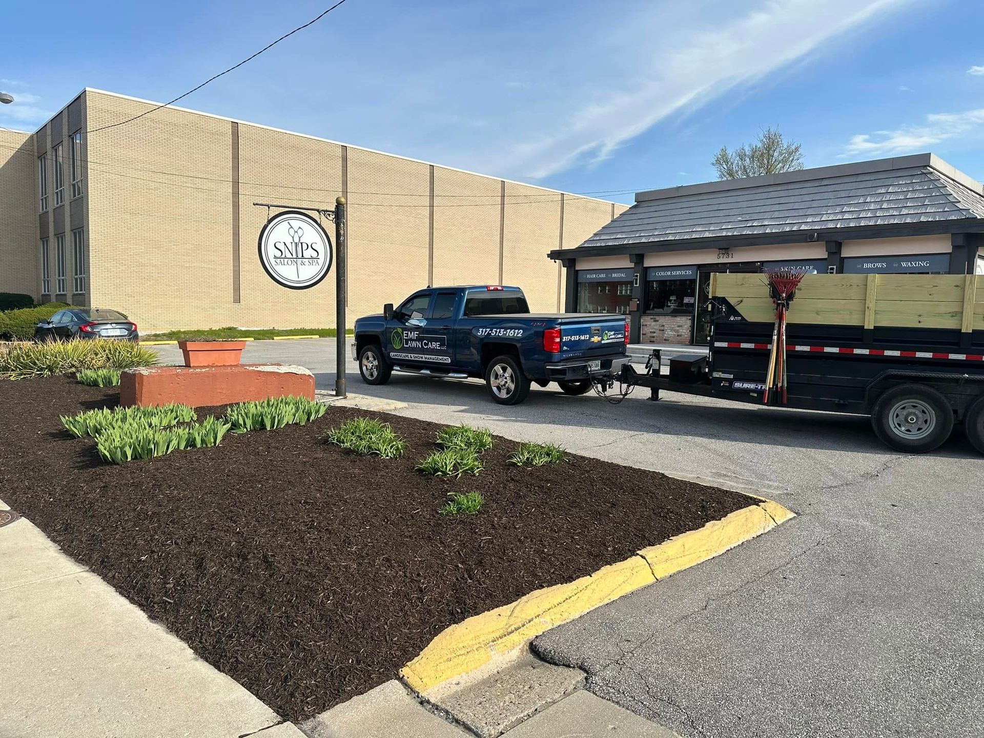 Outdoor clock on tan building beside parked pickup truck and trailer near a landscaped parking lot.