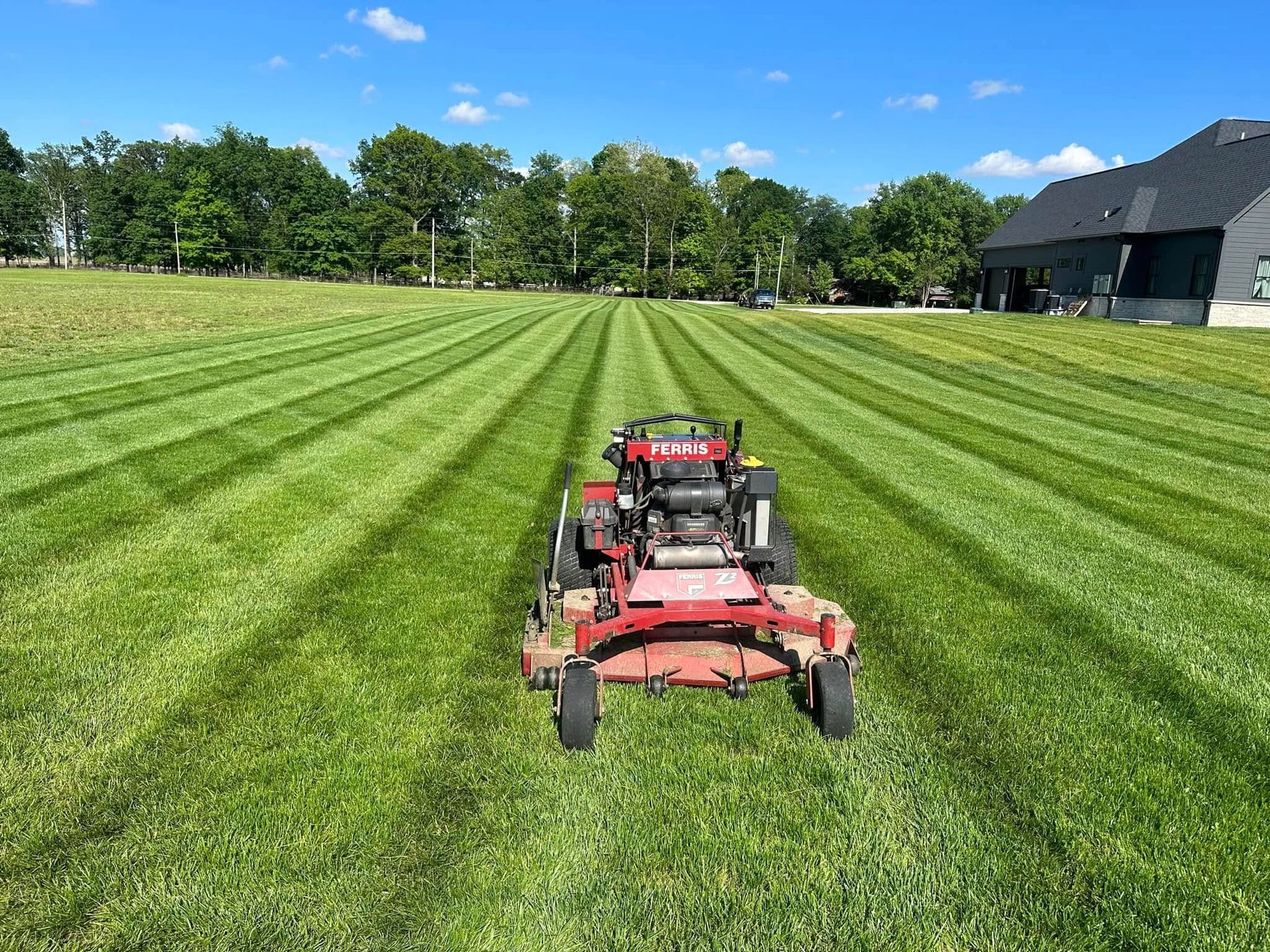 Red lawn mower on striped green field near trees and a barn under a clear blue sky