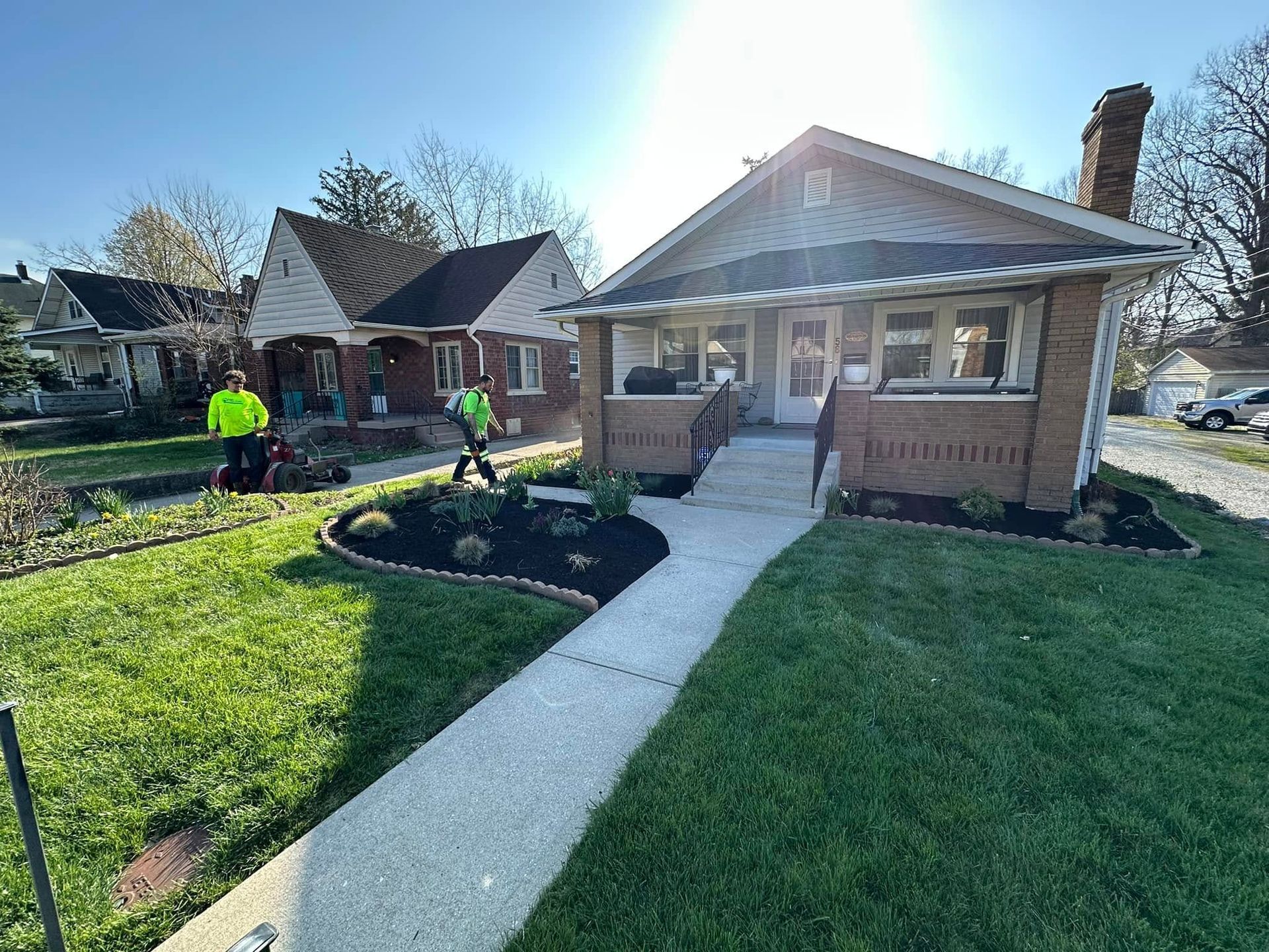 Brick bungalow with a front walkway, landscaped yard, and two workers in neon jackets outside.