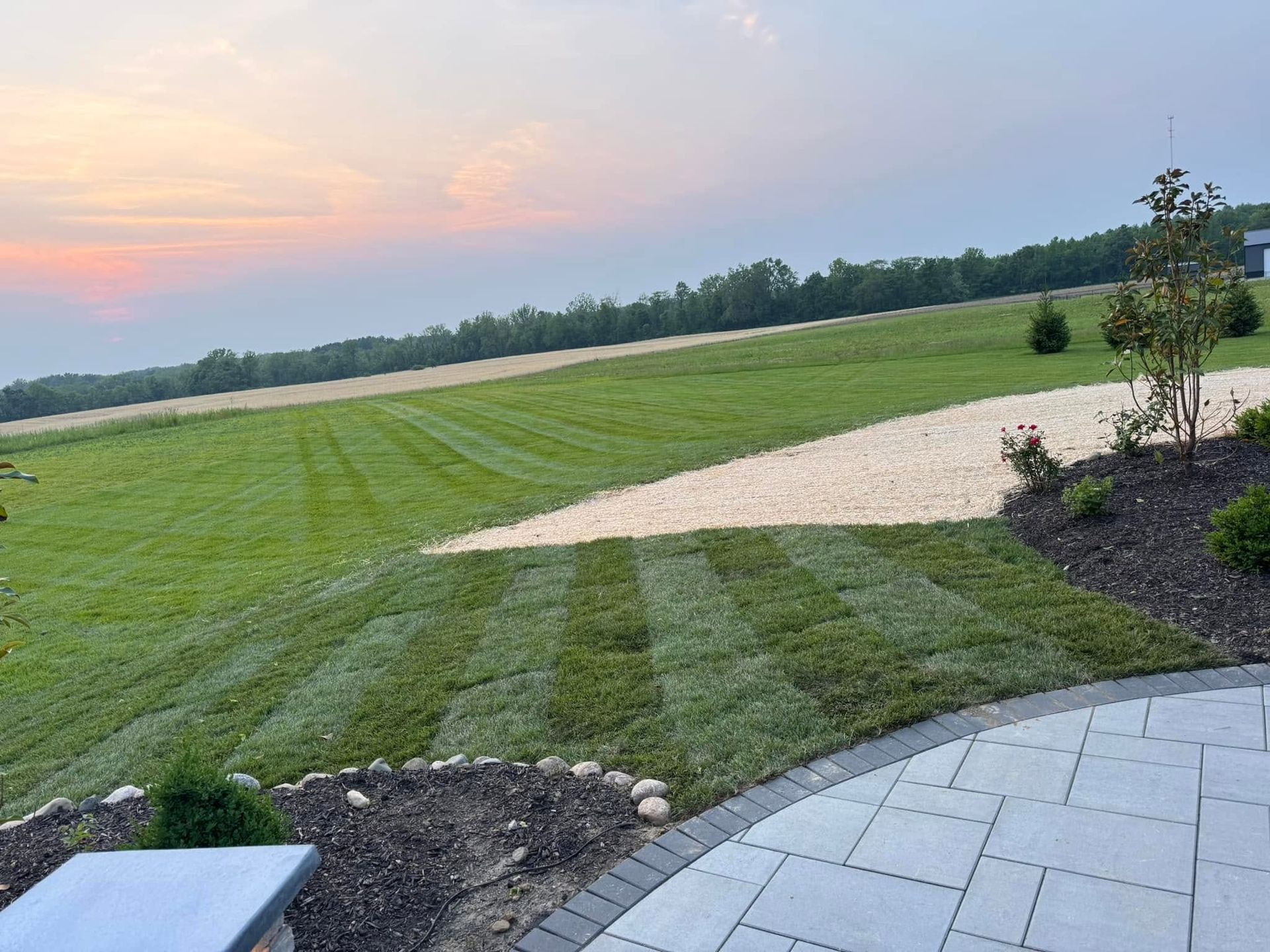 Curved stone patio overlooking a green lawn, gravel path, and sunset sky over open fields