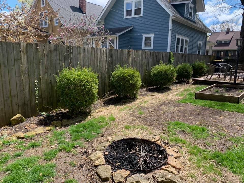 Backyard with a fire pit, wooden fence, trimmed shrubs, and a blue house in the background