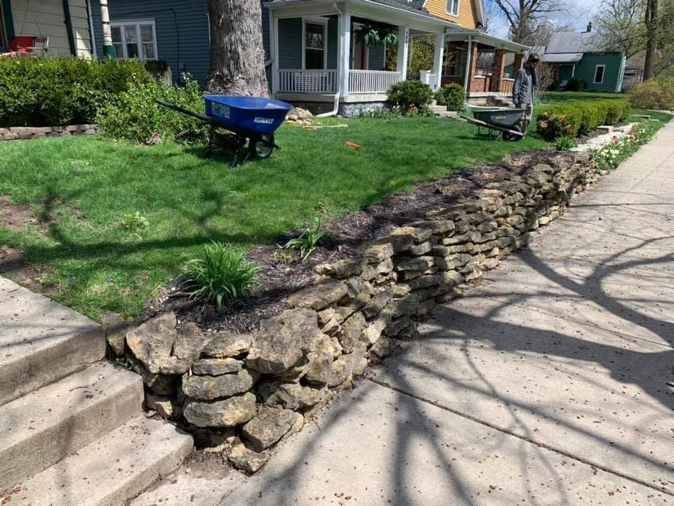 Stone retaining wall along a sidewalk beside a grassy yard and houses, with a blue wheelbarrow nearby.
