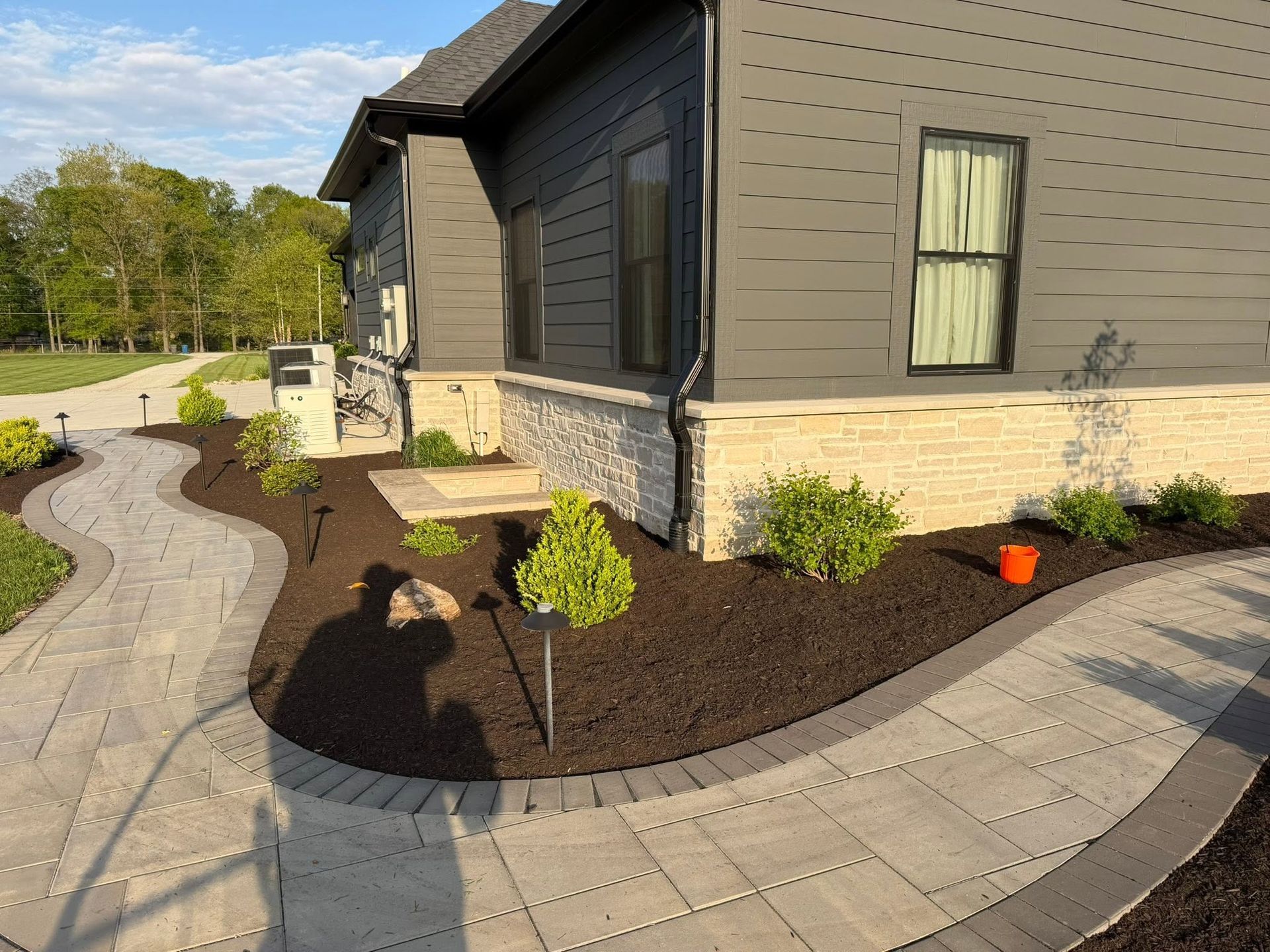 Curved paver walkway beside a gray house with fresh mulch, small shrubs, and a bright orange bucket.