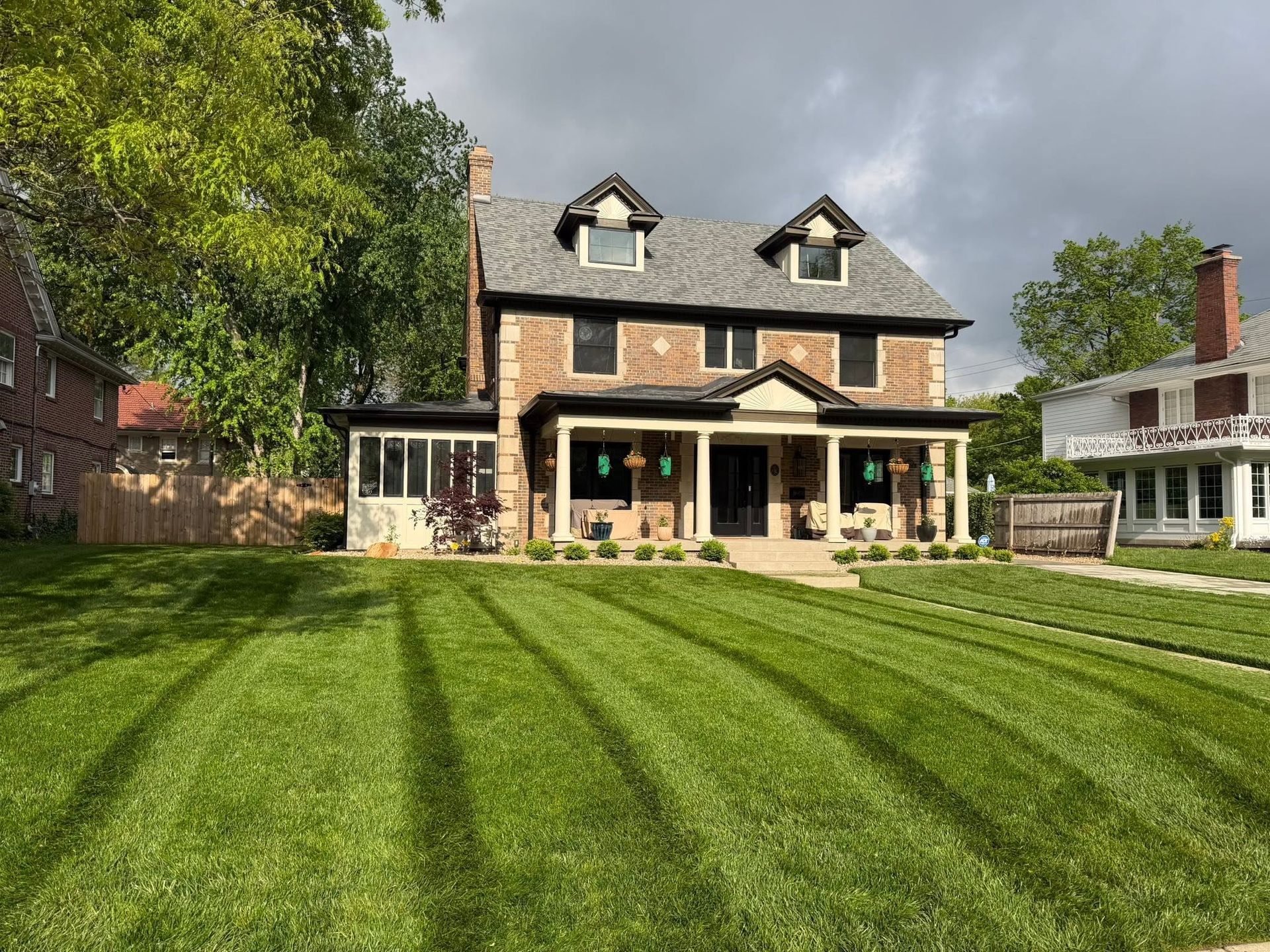 Two-story stone house with dormer windows and a wide striped green lawn under a cloudy sky
