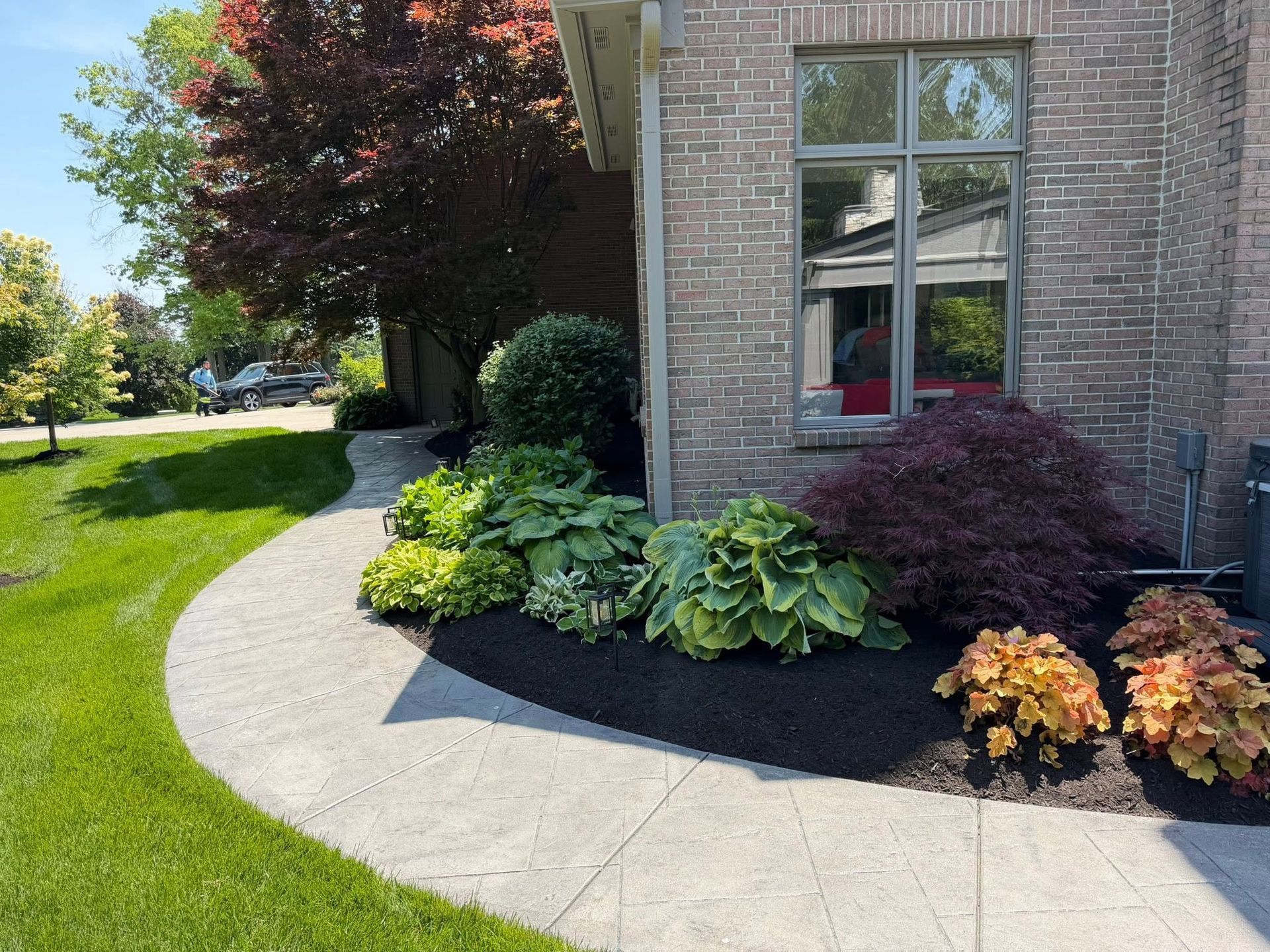 Curved concrete walkway beside a brick house with landscaped shrubs and flowers on a sunny day
