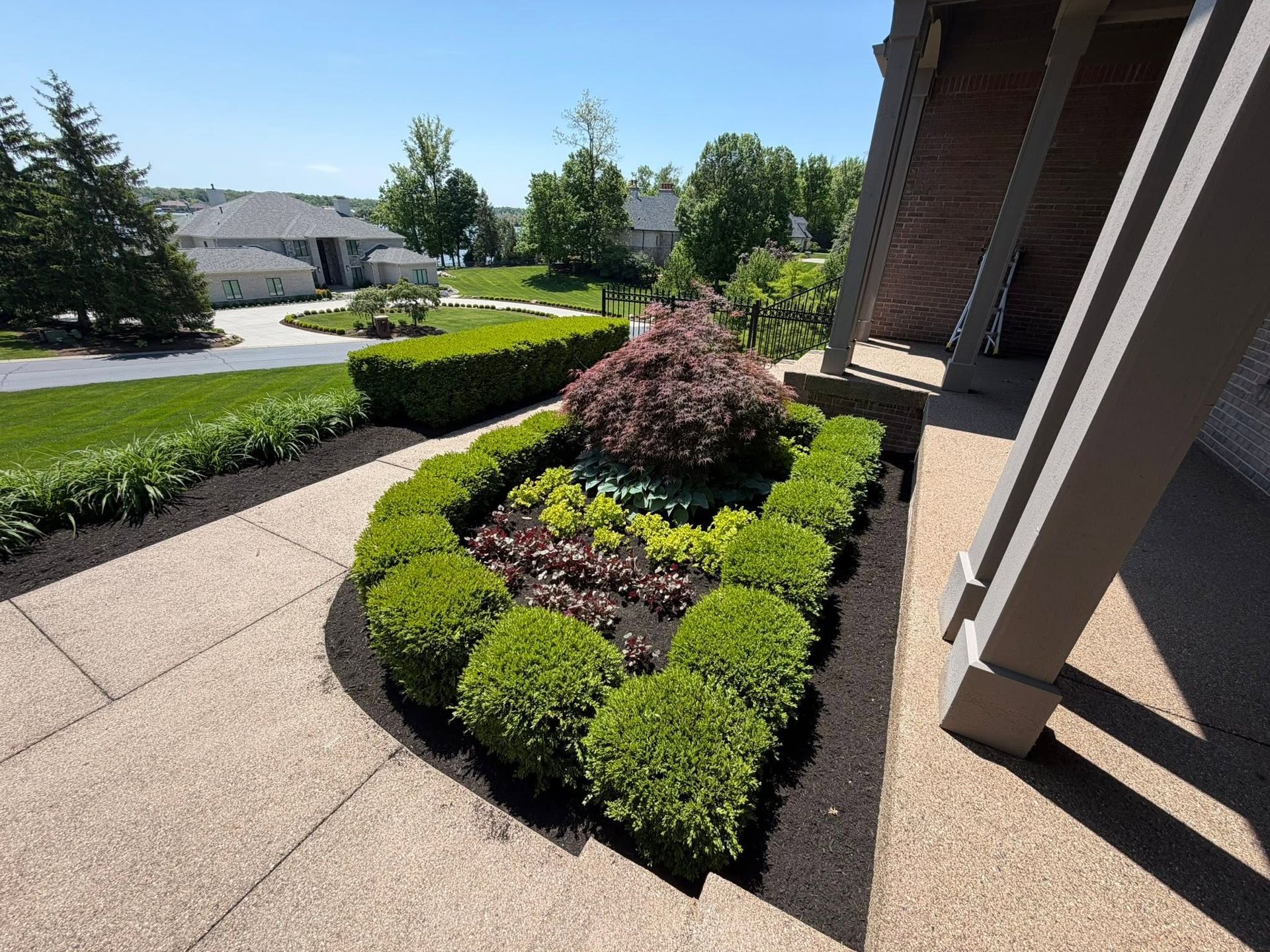 Trimmed front yard landscaping with green shrubs and a red bush along a house walkway on a sunny day