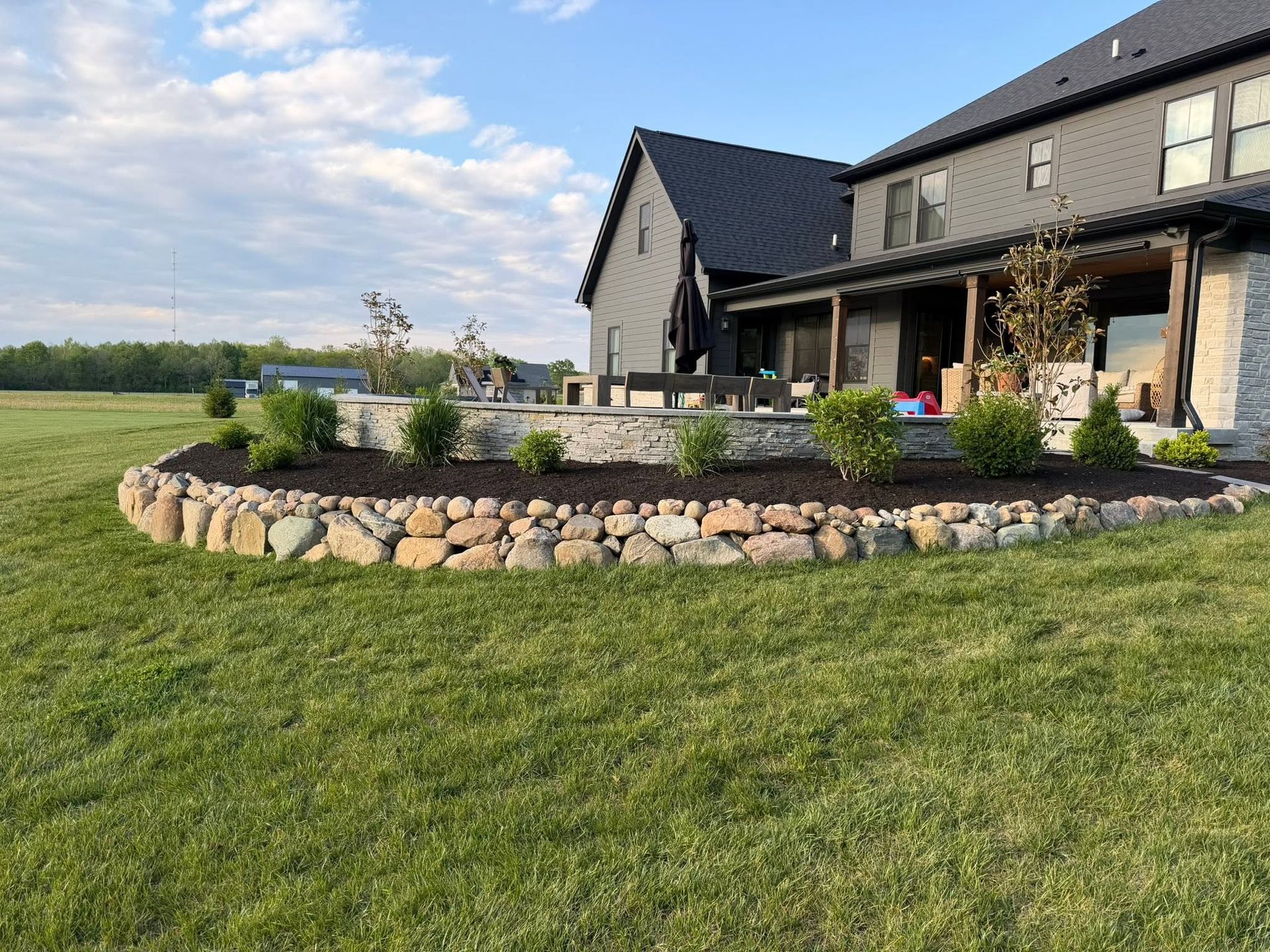 Stone-bordered landscaped flower bed beside a modern house with patio and green lawn under a blue sky