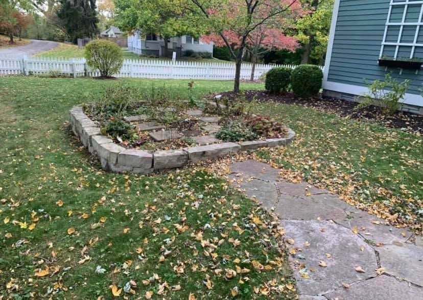 Stone garden bed beside a cracked walkway in a front yard with scattered autumn leaves and a white fence