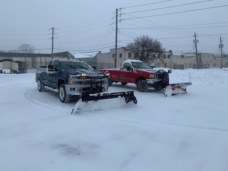 Two pickup trucks parked in a snowy lot, one black and one red, with snowplow attachments.