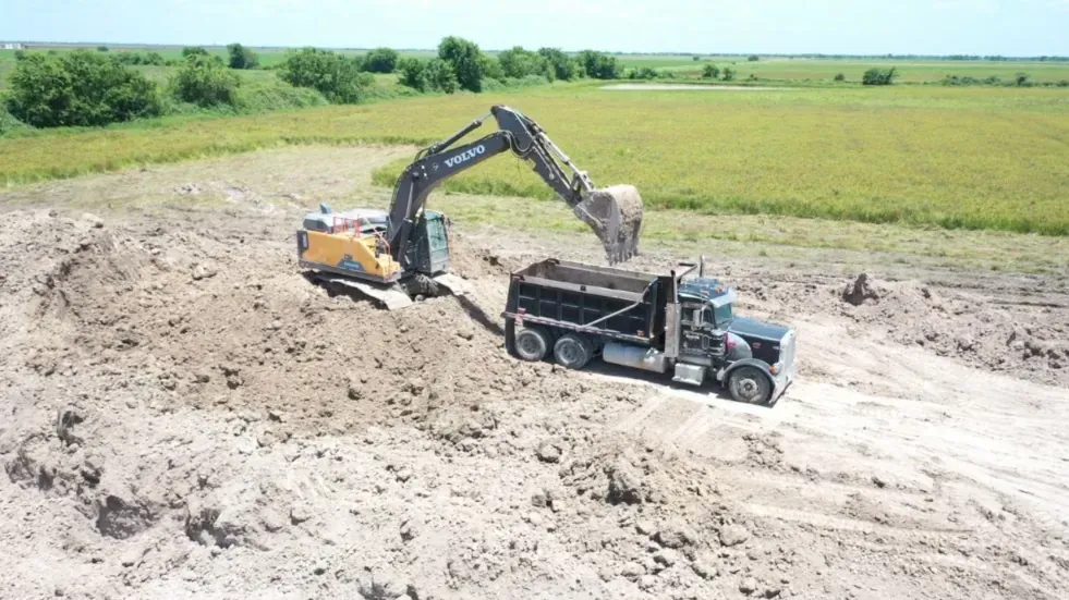 Excavator loading native pond soil into a tandem-axle dump truck for reuse onsite