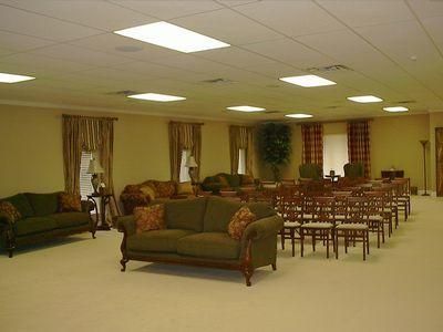 Spacious meeting room with green sofas, rows of chairs, curtains, and fluorescent ceiling lights.