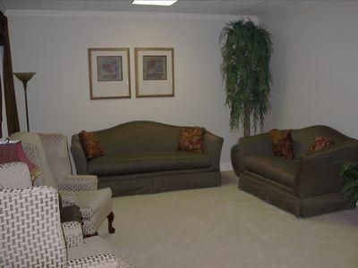 Living room with beige walls, framed art, dark sofas, a patterned chair, and tall potted plants