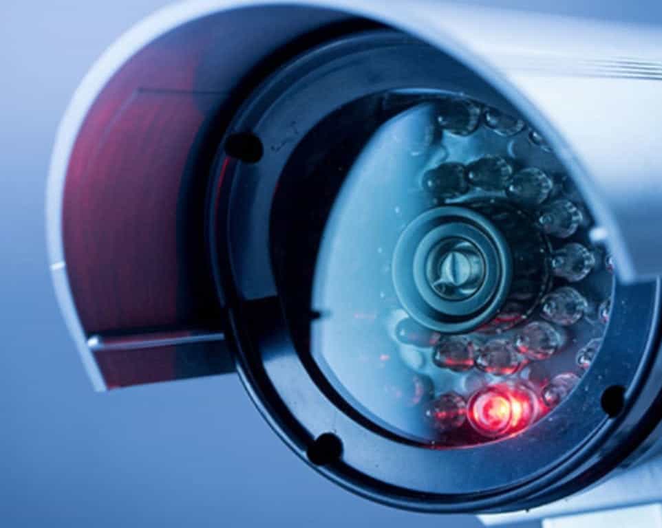 Security camera with red indicator light, close-up against a blue background.