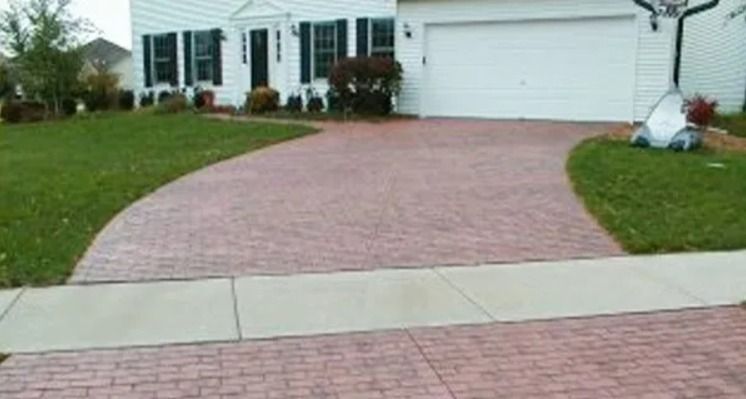 Brick-patterned reddish-brown driveway curving toward a white house with black shutters and a garage. Green grass surrounds.