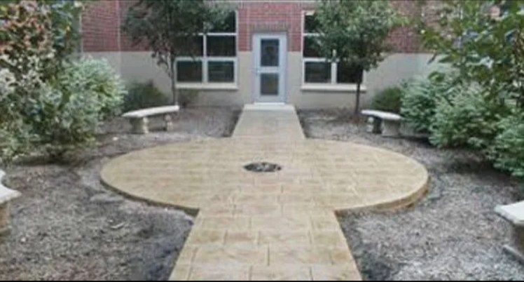 Concrete patio with a pathway leading to a building's door. Benches and landscaping are on either side.