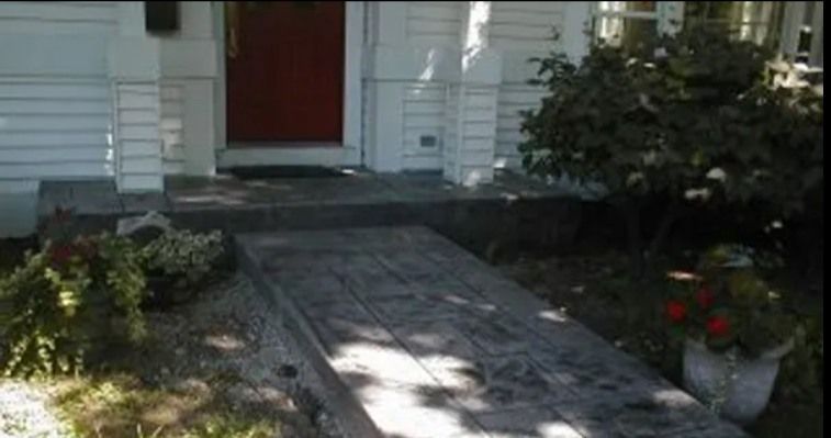 Wooden walkway leading to a house with a red door, flanked by greenery and bushes.