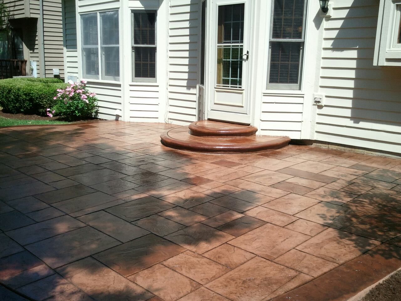 Brick-like patio in front of a white house with steps leading to a door; shadows cast on the ground.