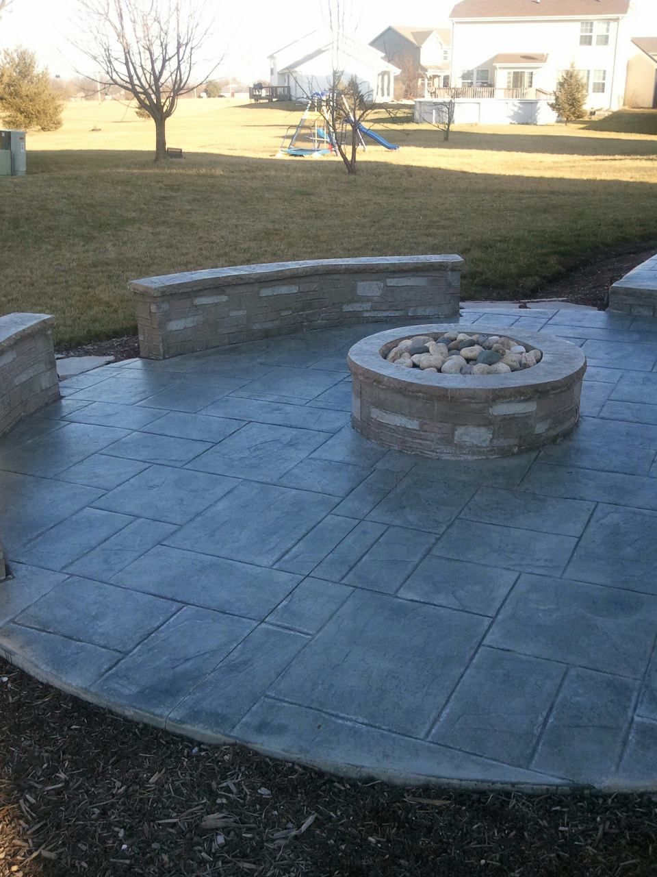 Stone patio with built-in fire pit, surrounded by low walls, in a backyard.