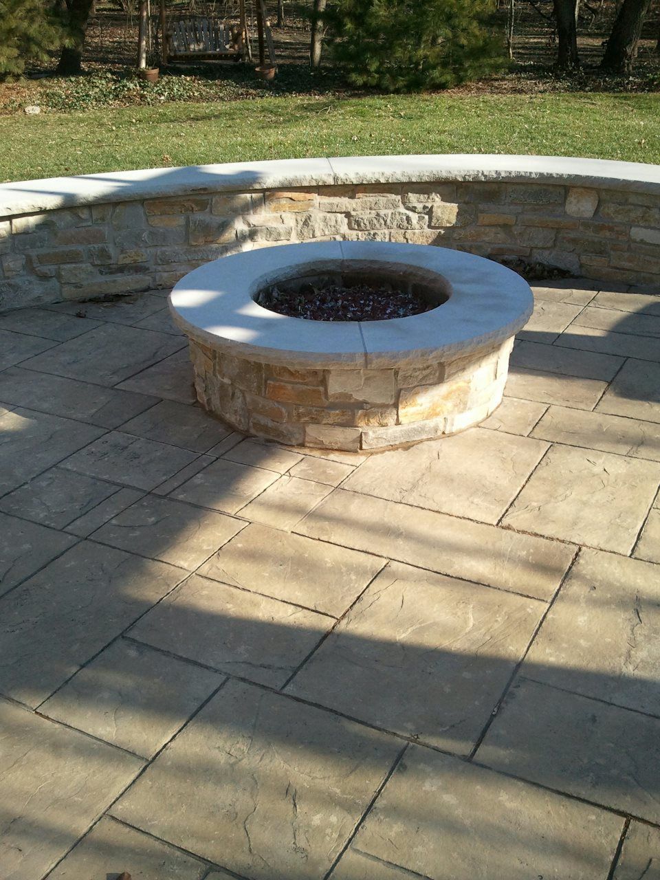 Circular stone fire pit on a stone patio, with a surrounding stone wall and green lawn in the background.