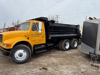 Yellow dump truck parked on gravel lot beside a black trailer