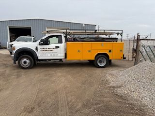 White utility truck with yellow service bed parked on a dirt lot beside a warehouse and gravel pile