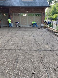 Three workers in high-visibility vests work on a grid pattern for a new concrete driveway in front of a brick garage.