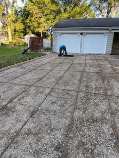 Person bending over on a cracked driveway in front of a two-car garage and trees.