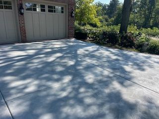 Sunlit concrete driveway beside a garage, with tree shadows and shrubs along the edge.