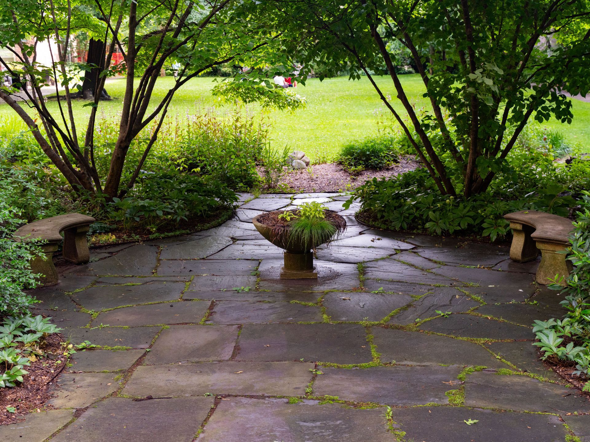 Stone garden patio with a small potted plant, framed by shrubs and trees opening to a green lawn.