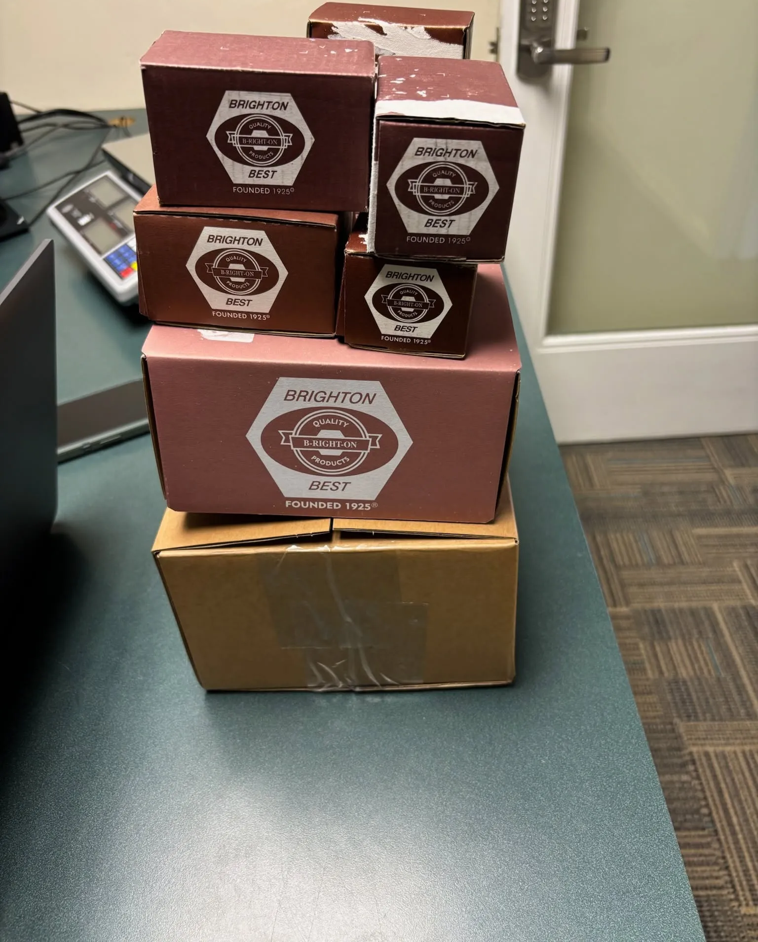 Stack of maroon-labeled boxes on a brown box in an office hallway beside a teal countertop.
