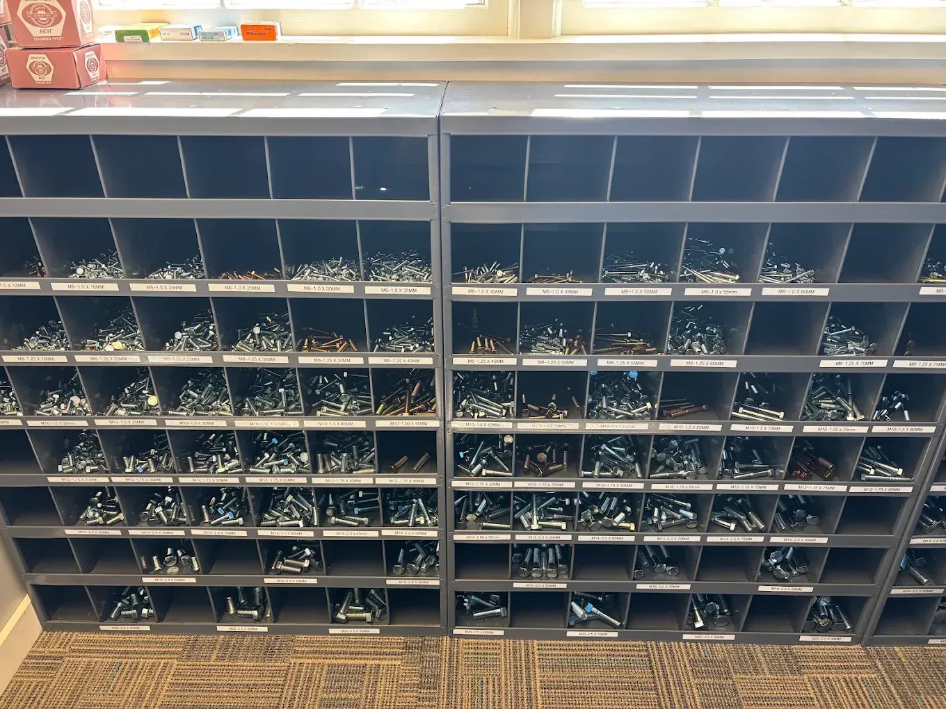 Rows of small parts bins on a wall shelf in a workshop, with metal hardware stored in compartments.