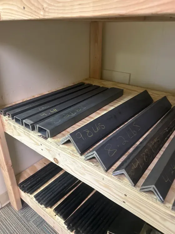 Shelves holding black metal V-shaped edge rails and trim pieces in a workshop storage area