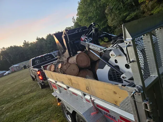 A utility trailer loaded with firewood and a piece of heavy machinery, attached to a pickup truck in a grassy field.