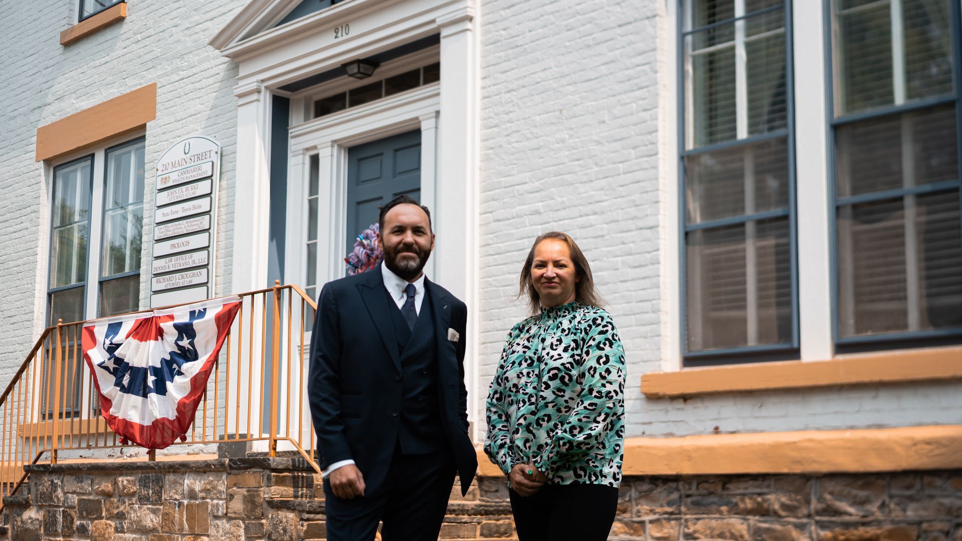 Two people stand outside a brick building with a patriotic banner. The man wears a suit, and the woman has a patterned shirt.