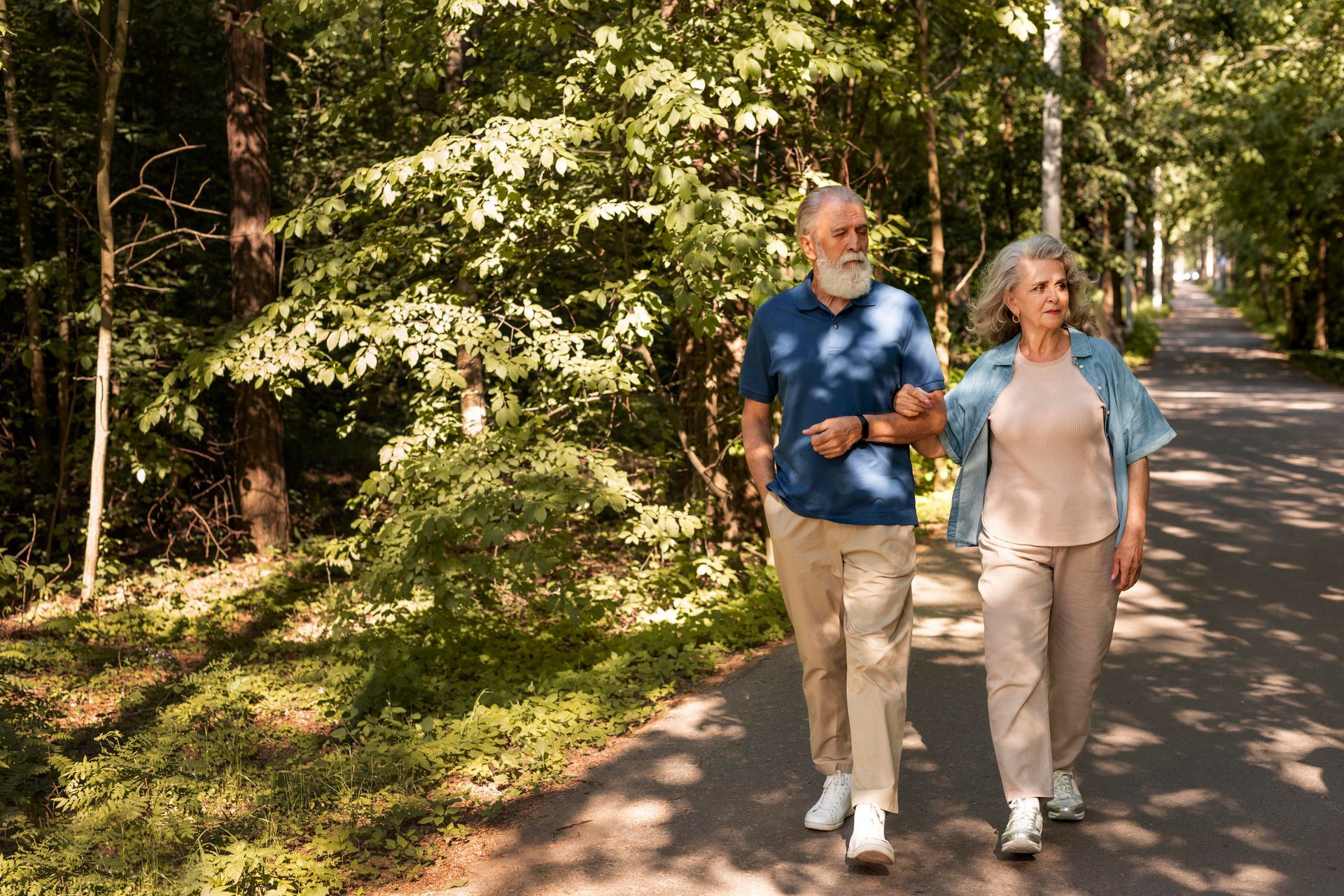 Older couple walking arm in arm on a path through a park, shaded by trees.