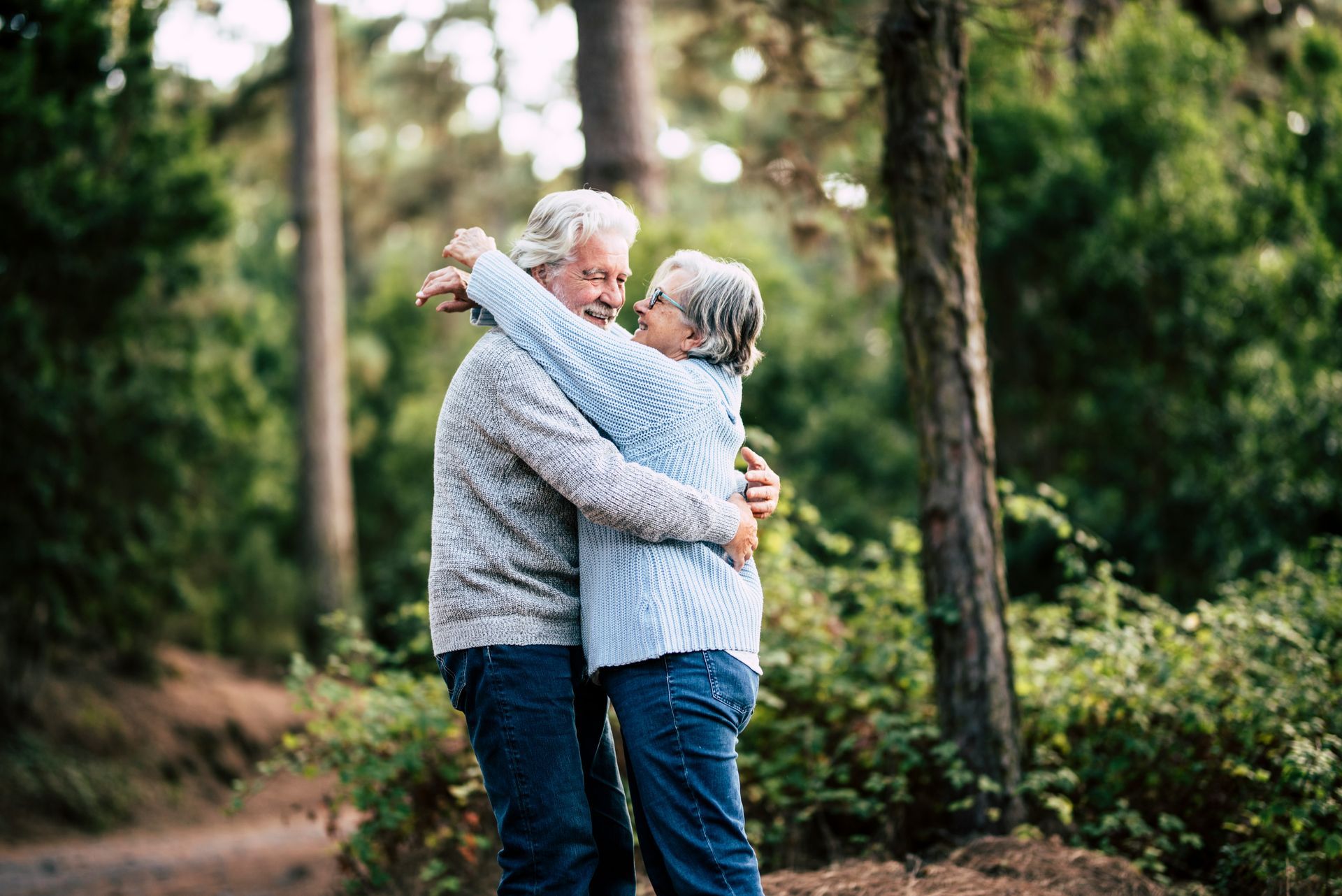 Older couple embracing in a forest; both smiling, wearing sweaters, surrounded by green trees.